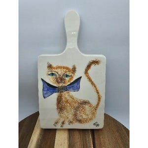 May include: White ceramic cutting board with a handle, featuring a hand-painted cat design. The cat has orange fur, blue eyes, and a blue bow tie. The board is rectangular with rounded corners.