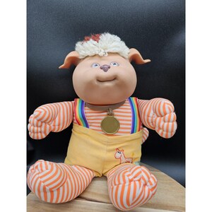 May include: A vintage plush toy featuring a dog-like character with a tan face, floppy ears, and a tuft of white and red hair. It wears orange and white striped arms and legs, yellow overalls, and a rainbow suspenders. A gold medallion hangs from its neck.