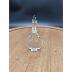 May include: A clear glass perfume bottle with a heart-shaped body featuring concentric embossed lines. The bottle has a pointed, teardrop-shaped glass stopper with the word CHAMADE etched in black lettering on the front surface of the stopper.