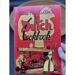 May include: A vintage cookbook titled "Edna Eby Heller's Dutch Cookbook" with a red cover. The cover features a stylized illustration of a Dutch kitchen scene and the word "Dutch" in large, bold letters. The book is labeled "Revised".