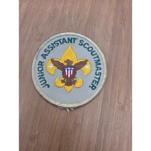 May include: A circular embroidered patch with a light gray background and a tan border. The text JUNIOR ASSISTANT SCOUTMASTER is stitched in dark green. In the center, a yellow fleur-de-lis emblem features a brown eagle with a red, white, and blue shield.