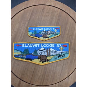 May include: Two embroidered patches featuring a blue sky, mountains, a lake, and a brown bear. The text ELAUWIT LODGE 37 is stitched in white, with red arrows on the sides and a yellow fleur-de-lis symbol. The patches have a yellow border.