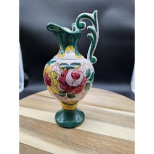 May include: A small, decorative ceramic pitcher with a floral design. The pitcher features a green handle and spout, with a base and accents in green and yellow. The body of the pitcher is decorated with red and yellow flowers. The pitcher is approximately 6 inches tall.