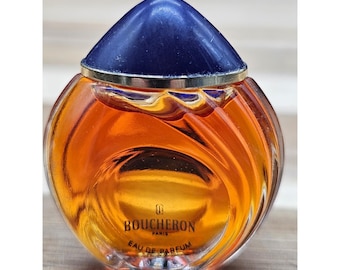 May include: A round glass perfume bottle filled with amber liquid, featuring a dark blue dome-shaped cap and a gold-tone band. The front of the bottle displays the text BOUCHERON PARIS EAU DE PARFUM with a small logo above the brand name.