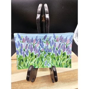 May include: Rectangular glass dish with a floral design. The artwork features lavender and pink flowers with green leaves against a light blue background. The dish is supported by a dark brown stand, and is displayed on a wooden surface.