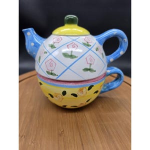 May include: A ceramic tea for one set featuring a teapot stacked on a teacup. The teapot has a white body with a blue diamond grid pattern, pink rose motifs, and a yellow lid. The teacup is yellow with painted yellow roses and a blue handle.