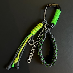 Könnte beinhalten: Ein schwarzer Karabinerhaken mit neongrünen und grauen Paracord-Akzenten. Der Haken hält eine kleine Kette, eine Handschlaufe und ein Schlüsselband, alle im gleichen Farbschema. Die Artikel werden vor einem schwarzen Hintergrund präsentiert.