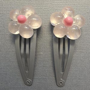 May include: Two silver barrettes, each adorned with a flower design. The flowers feature translucent petals and a pink center. The barrettes are set against a light blue background.