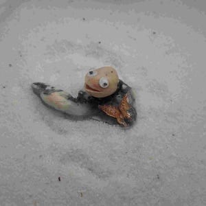 May include: A whimsical, handmade turtle figurine with a light tan head, large googly eyes, and a smiling mouth. The turtle's body is a mix of gray, white, and tan, with a small, orange-brown butterfly attached. The figurine is set against a white background.