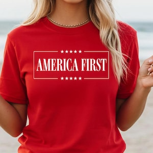 Peut inclure: Un t-shirt rouge avec le texte "AMERICA FIRST" dans un rectangle blanc, avec des étoiles au-dessus et en dessous du texte. La personne porte un short blanc et tient des drapeaux américains.