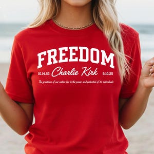 May include: A red t-shirt with the word "FREEDOM" in white block letters. Below the word "FREEDOM" is the name "Charlie Kirk" and the dates "10.14.93" and "9.10.25". The text "The greatness of our nation lies in the power and potential of its individuals" is also on the shirt.