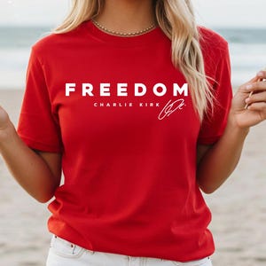 May include: A red t-shirt with the word "FREEDOM" in white block letters. Below the word "FREEDOM" is the text "CHARLIE KIRK" and a signature. The person is holding two American flags.