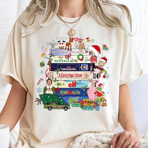 Puede incluir: Camiseta color crema con una pila de cintas VHS de películas navideñas con ilustraciones festivas y títulos como "Solo en casa" y "Elf". El diseño incluye personajes y decoraciones navideñas, creando un tema de temporada.