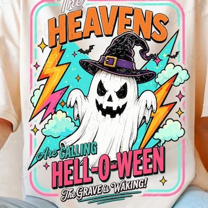 May include: A cream-colored t-shirt featuring a graphic of a ghost wearing a purple witch hat. The design includes orange lightning bolts, blue clouds, and the text: The HEAVENS Are CALLING HELL-O-WEEN The GRAVE is WAKING! The print is framed by a colorful border.
