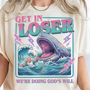 May include: Cream-colored t-shirt with a graphic design featuring a whale with an open mouth, waves, and a man in the water. Text reads "GET IN LOSER" and "WE'RE DOING GOD'S WILL" in a retro style.