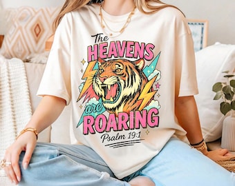 May include: A cream-colored short-sleeve t-shirt featuring a graphic of a roaring tiger head surrounded by pink lightning bolts and blue clouds. The text reads The HEAVENS are ROARING Psalm 19:1 in bold pink and black lettering on the front.