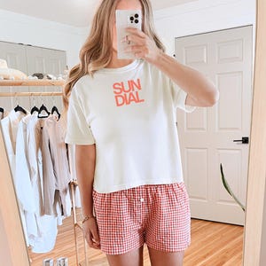 May include: Cream-colored t-shirt with the word "SUN DIAL" in red block letters. Paired with red and white checkered shorts. The outfit is reflected in a mirror.