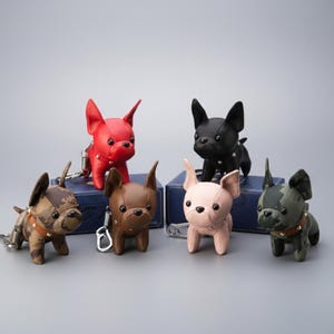 May include: A collection of six French bulldog keychains in various colors, including red, black, brown, pink, and camouflage patterns. Each keychain features a detailed dog design with a metal clasp for attaching to keys or bags.