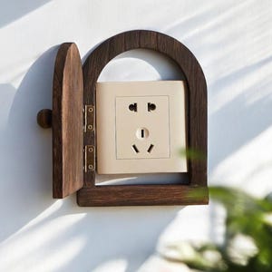 May include: A decorative wooden outlet cover with a hinged door. The cover is dark brown and surrounds a beige electrical outlet. The door is open, revealing the outlet. The design is mounted on a white wall.