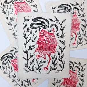 May include: A collection of red and black linocut prints featuring a whimsical house with long, bird-like legs, a smoking chimney, and surrounded by leafy vines. The house has a detailed facade and is printed on textured paper.