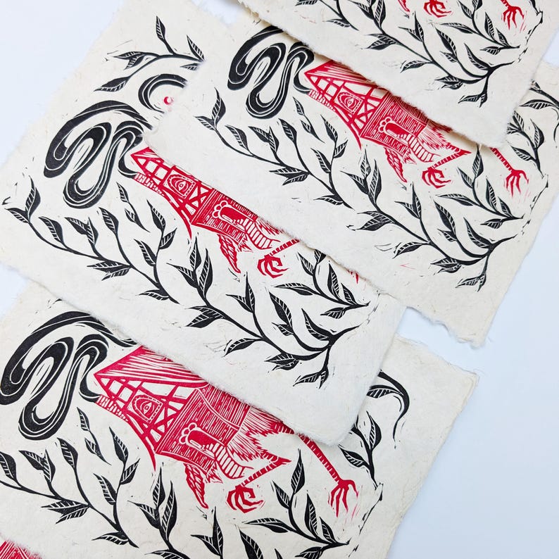May include: A collection of linocut prints featuring a red and black design. The prints showcase a stylized creature with a house-like form, intertwined with leafy branches and swirling black shapes. The prints are on textured, off-white paper.