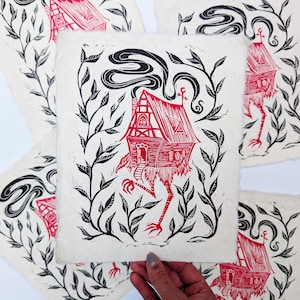 May include: Red and black linocut print featuring a whimsical house with chicken legs, surrounded by black foliage. The house has a detailed facade and smoke billowing from the chimney. The artwork is on textured, off-white paper.