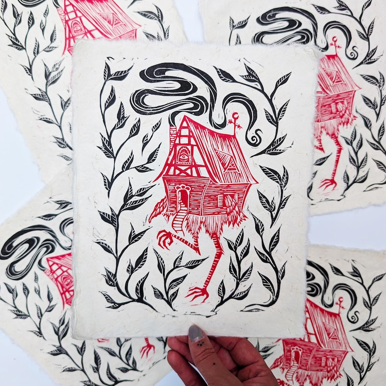 May include: Red and black linocut print featuring a whimsical house with chicken legs, surrounded by black foliage. The house has a detailed facade and smoke billowing from the chimney. The artwork is on textured, off-white paper.