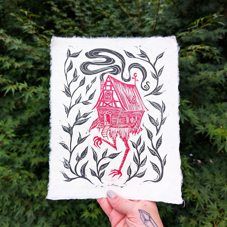 May include: A red and black linocut print of a whimsical house with legs, surrounded by leafy vines. The house has a chimney with smoke and a decorative roof. The artwork is on textured, off-white paper.