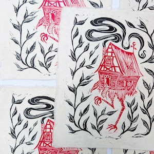 May include: Red and black linocut prints featuring a whimsical house with chicken legs, surrounded by black leafy vines. The house has a chimney with smoke and an eye on the front. The prints are on textured, off-white paper.