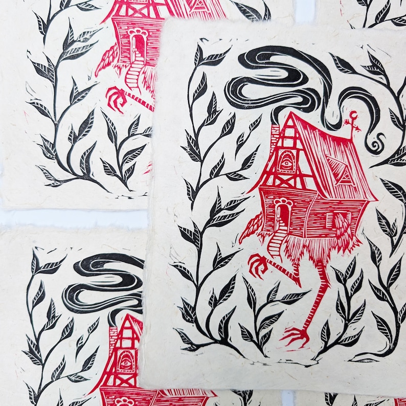 May include: Red and black linocut prints featuring a whimsical house with chicken legs, surrounded by black leafy vines. The house has a chimney with smoke and an eye on the front. The prints are on textured, off-white paper.