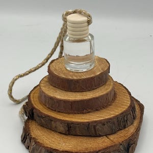 Puede incluir: Una botella de vidrio transparente con una tapa de madera y una cuerda colgando. La botella se encuentra sobre una base de madera escalonada hecha de rodajas de madera circulares. La madera es de color marrón natural.