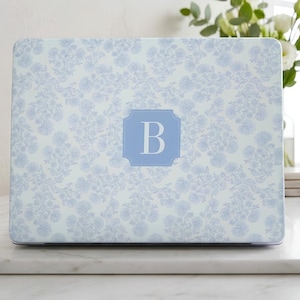 May include: A laptop skin featuring a light blue and white floral toile pattern. In the center, a rectangular blue badge with clipped corners displays the white capital letter B. The design covers the entire surface of the closed laptop lid.