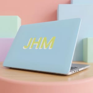 May include: A light blue laptop with the letters JHM printed in a yellow font with a white shadow effect on the lid. The laptop is positioned at an angle, displaying the screen cover and the side profile with visible ports and a metallic edge.