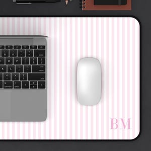 May include: A rectangular desk mat featuring a light pink and white vertical striped pattern with the letters BM printed in the bottom right corner. The mat provides a surface for a silver laptop and a white computer mouse.