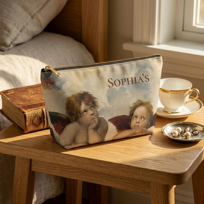 May include: A beige cosmetic bag with a gold zipper, featuring a Renaissance-style painting of two cherubs against a cloudy sky. The name "SOPHIA'S" is printed on the bag. A teacup and saucer are on a small table.
