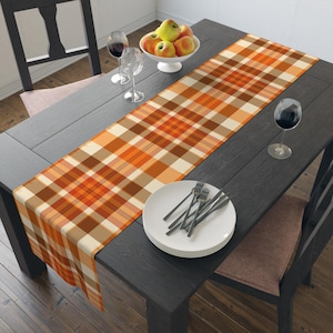 May include: A rectangular table runner featuring a plaid pattern in shades of orange, brown, and cream. The runner is laid across a dark wooden table set with a stack of white plates, silver forks, two wine glasses, and a bowl of apples.