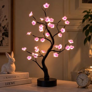 May include: A decorative LED tree lamp featuring a dark brown trunk and branches adorned with numerous small, glowing pink cherry blossom-shaped lights. The tree is mounted on a circular black base and emits a soft, warm light from each flower.