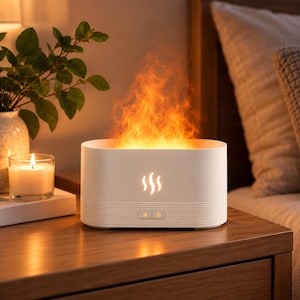 May include: A white oval-shaped essential oil diffuser featuring a flame-like light effect emitting from the top. The front of the device displays a glowing flame icon and two control buttons, with horizontal ribbed detailing along the base.