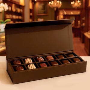 Peut inclure: Une boîte cadeau rectangulaire marron avec un couvercle à charnière, remplie d'un assortiment de chocolats. Les chocolats sont de formes et de couleurs variées, notamment marron clair et foncé. La boîte est ouverte, révélant les friandises à l'intérieur.