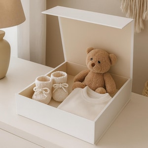 Pode incluir: Caixa de presente branca aberta com um ursinho de pelúcia castanho, uma camiseta branca e botinhas de bebê creme com laços. O conjunto é apresentado em uma caixa. Tema: presentes para bebês.
