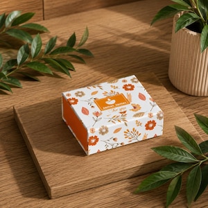 May include: A square gift box with a white background featuring an orange and brown floral pattern. The center of the lid displays an orange rectangular label with the white text James James For you and a small icon of a lit candle.