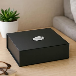 Può includere: Una scatola quadrata nera con un logo a forma di nuvola bianca. La scatola è appoggiata su una superficie di legno chiaro, accanto a una piccola pianta succulenta in vaso. Il testo sulla scatola recita "cloud design".