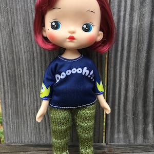Puede incluir: Una muñeca con cabello rojo que lleva una camisa azul con el texto "DOOOOOH!" y pantalones verdes. La muñeca tiene ojos azules y mejillas sonrosadas.