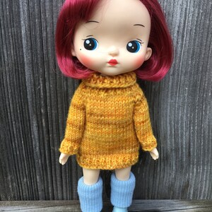 Puede incluir: Una muñeca con ojos azules brillantes, cabello rojo y un suéter de punto amarillo. La muñeca lleva polainas azules y zapatos azules.