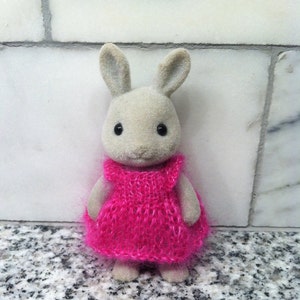 Puede incluir: Un pequeño conejo gris con un vestido de punto rosa. El conejo está de pie sobre una superficie de azulejos grises y blancos.