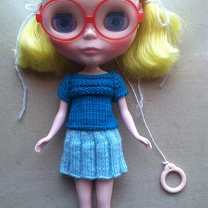 May include: A doll with blonde hair wearing a blue knitted sweater and a light blue knitted skirt. The doll is wearing red glasses and has a white string attached to a ring.