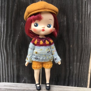 Puede incluir: Una muñeca con el pelo rojo que lleva un jersey azul y amarillo, un sombrero marrón y pantalones cortos amarillos. La muñeca tiene ojos azules y una boca roja.