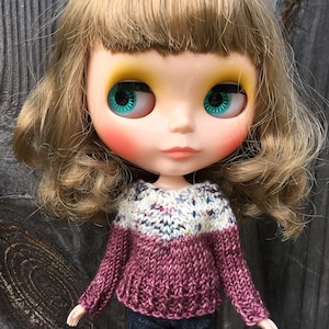 May include: A doll with long brown hair wearing a pink and white knitted sweater. The doll has blue eyes and rosy cheeks.