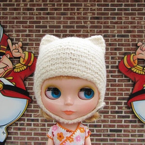 Op de afbeelding: Een witte gebreide muts met kattenoren, gedragen door een pop met blauwe ogen en een bloemenjurk. De pop staat voor een bakstenen muur met twee cartoonfiguren aan weerszijden.
