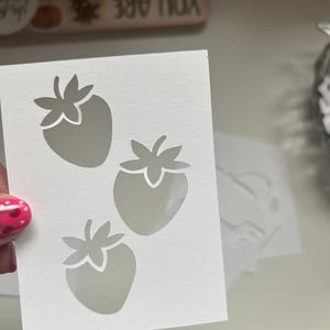 May include: A white rectangular stencil featuring three die-cut strawberry shapes arranged in a vertical pattern. Each strawberry cutout includes a detailed leafy stem at the top, creating a clean and simple design for crafting and artistic projects.